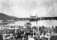 Spectators mill around the bandstand at the 1905 Lewis and Clark  Exposition, with the lake and the Government Building behind. The largest part of the lake is behind the peninsula the bridge and peninsula the building is on. (Robert A. Reid, Portland)