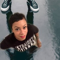 Artist Try Cheatham looking up at the camera whilst standing on a reflective surface. Someone stands behind her with only their black-and-white canvas shoes shoes in the camera frame.
