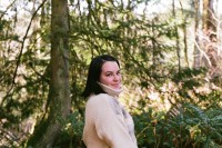 Photo of Zowie in the woods. They have short to medium length dark hair and a white, wool coat on that comes up to their chin. She is surrounded by cedar trees and ferns.