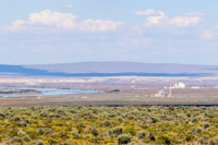 Panoramic view of Hanford Nuclear Reservation. There is a blue sky with whispy clouds in the distance. There are blue-ish brown low mountains in the background and in the foreground the iconic green sage brush shrubs fill the photo