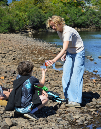 Photo of Jenna handing a water quality sample to a child who has their back turned from the camera. They are standing on rocks along the river from and there are trees in the background. You can see the edge of the water peeking from behind Jenna.