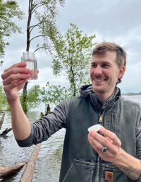 Theus, our water quality intern, holding up a water sample. He is smiling and looking at the container. There are trees in the background and cloudy skies.