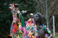 Image of the statue of "origami dreams of Sadako Sasaki" who is covered by hundreds of colorful origami cranes. There are trees in the background, 