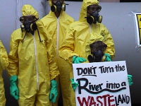 people protesting in hazmat suits with a sign that says "don't turn the river into a wasteland"