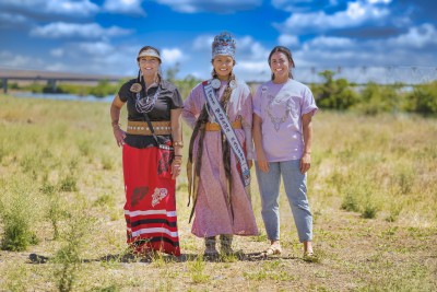 Three women standing in a grassy, prairie landscape with a blue sky and clouds in the background.