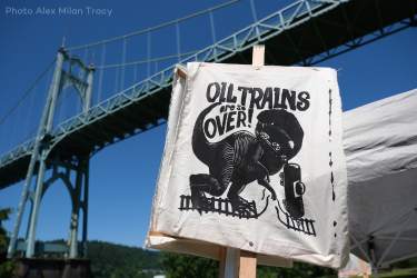 Image of poster and in the background is a green bridge and sky, Poster reads "Oil trains are over!" with a dinosaur ripping up an oil train. Taken at the Zenith Climate Rally in summer of 2023.