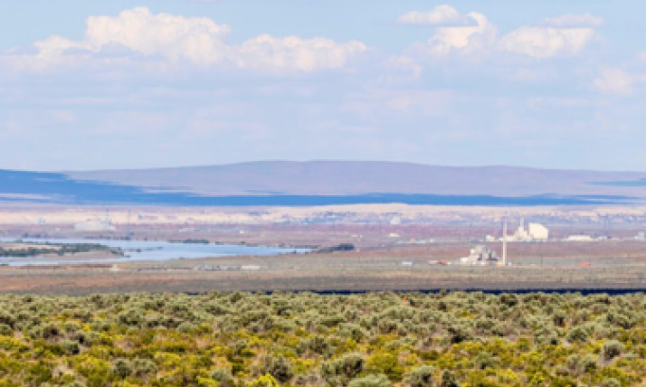 Panoramic view of Hanford Nuclear Reservation. There is a blue sky with whispy clouds in the distance. There are blue-ish brown low mountains in the background and in the foreground the iconic green sage brush shrubs fill the photo