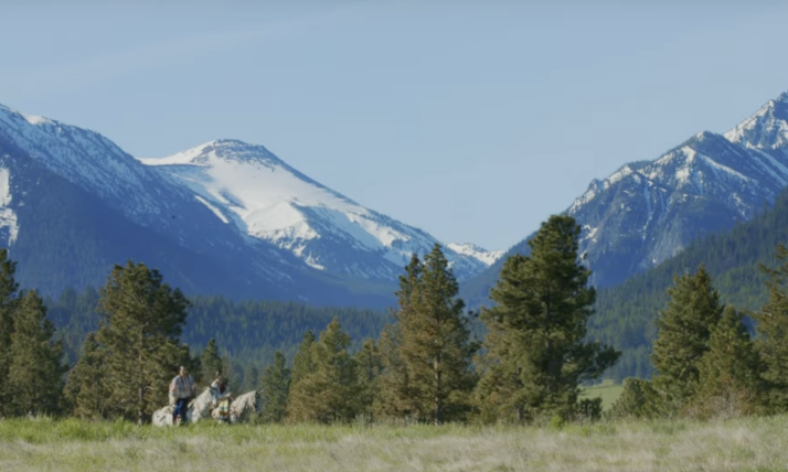 Image from film shows mountains in the background with green trees and grass in for foreground. Very small, you can see two people on horses approaching from the left corner. Image credit to film makers. 