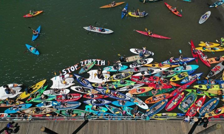 In June 2023, activists gathered in Portland at the Land and Water Rally Day of Action against fossil fuels. Aerial image of a dock and water with multicolors kayaks in the water and banner that says "Stop Zenith"