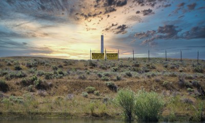 Photo of the Hanford Reach showing water, a hill with grass, and a beautiful sunset contrasting with a sign that reads "Warning: Hazardous area, do not enter" and fence.